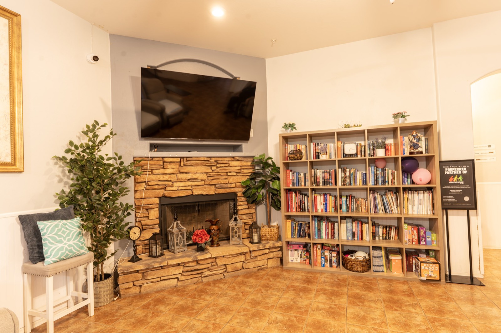 Cozy common room with a stone fireplace, plants, and a shelf of books and games